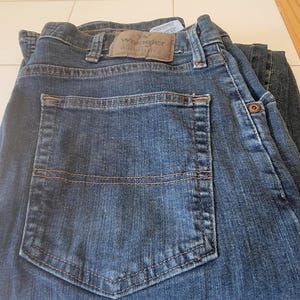 May include: Dark blue denim jeans with a classic five-pocket design. The jeans feature a brown leather patch with the "Wrangler Authentics" logo. The back pocket has a stitched design. The jeans are folded.