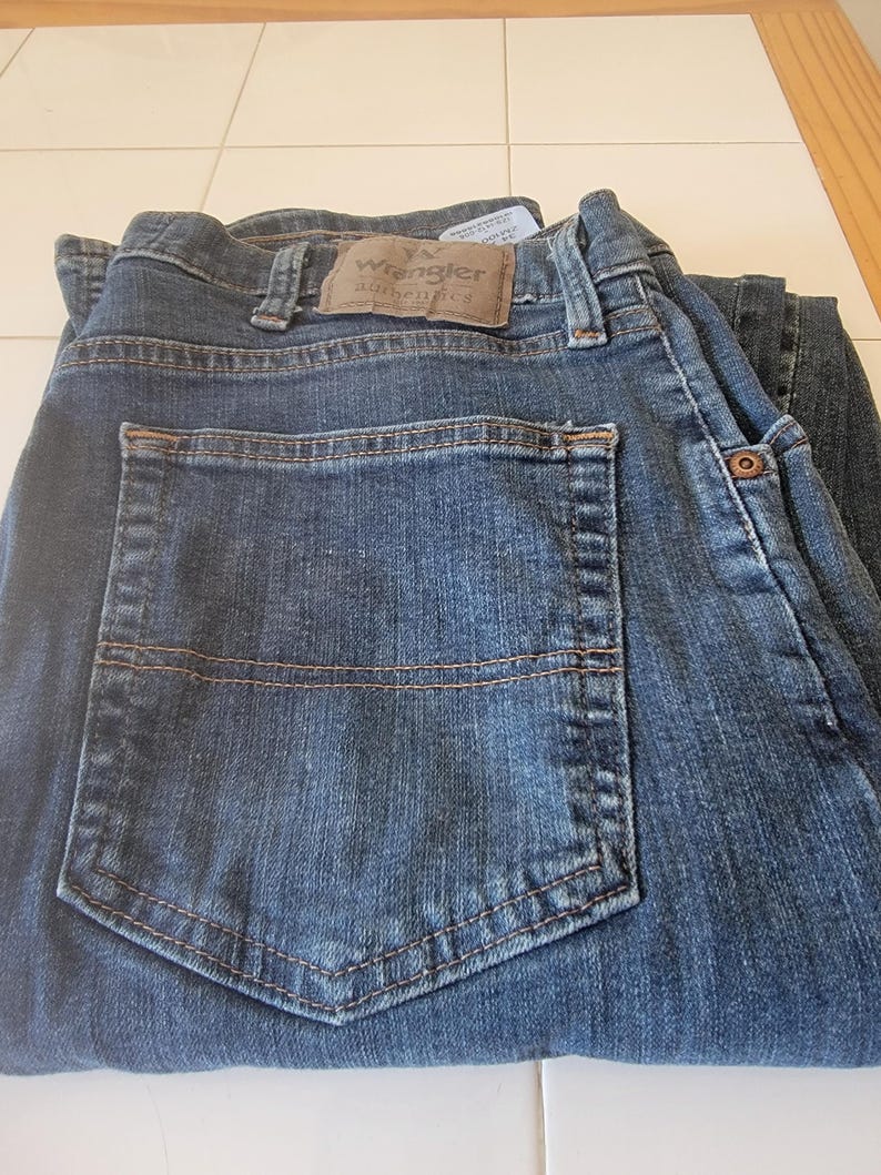 May include: Dark blue denim jeans with a classic five-pocket design. The jeans feature a brown leather patch with the "Wrangler Authentics" logo. The back pocket has a stitched design. The jeans are folded.