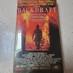 May include: A VHS tape case for the movie Backdraft, featuring a firefighter walking through a fiery scene. The case has a red background with the movie title in white letters. The actors Kurt Russell, William Baldwin, Scott Glenn, Jennifer Jason Leigh, Rebecca De Mornay, Donald Sutherland, and Robert De Niro are listed on the front of the case.