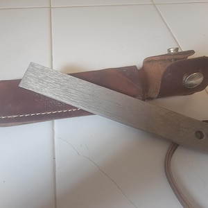 May include: A rectangular metal sharpening stone with a brushed finish and a small hole at one end, accompanied by a dark brown leather sheath featuring a metal snap button closure and visible stitching along the edges of the leather material.