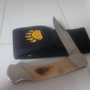 May include: A folding pocket knife with a silver blade marked LB-5 and a cream-colored handle with brown mottled accents. The knife is positioned in front of a black nylon sheath featuring an embroidered yellow bear paw print design.
