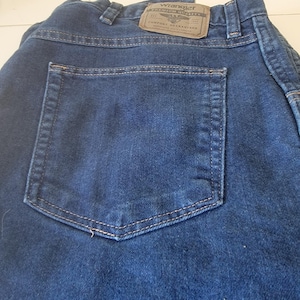 May include: Close-up of dark blue denim jeans with a back pocket. The jeans have a brown leather patch with the "Wrangler" logo and the text "Premium Quality Comfort Guaranteed". The stitching is a light tan color.
