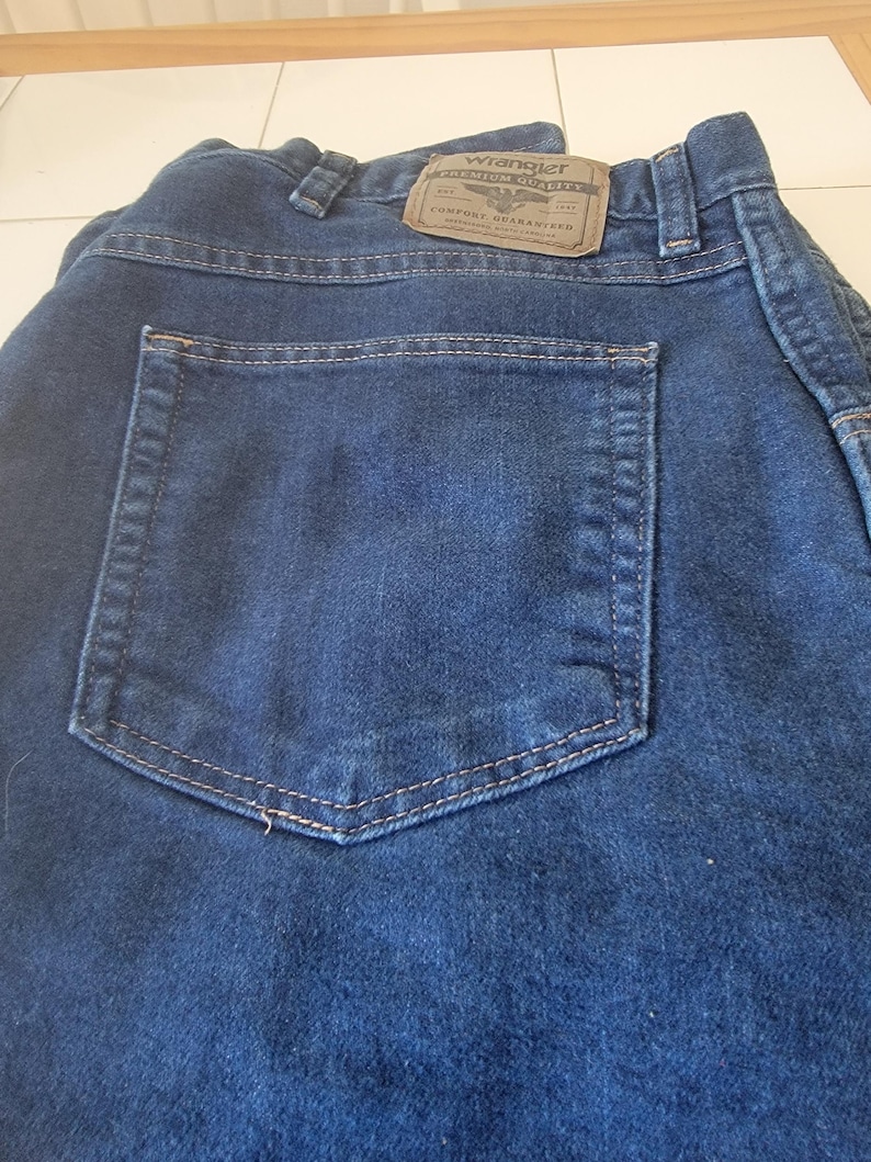 May include: Close-up of dark blue denim jeans with a back pocket. The jeans have a brown leather patch with the "Wrangler" logo and the text "Premium Quality Comfort Guaranteed". The stitching is a light tan color.