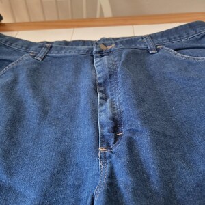May include: Close-up of blue denim jeans. The jeans have a button closure and pockets. The stitching is visible in a contrasting color. The fabric appears to be a medium weight denim.