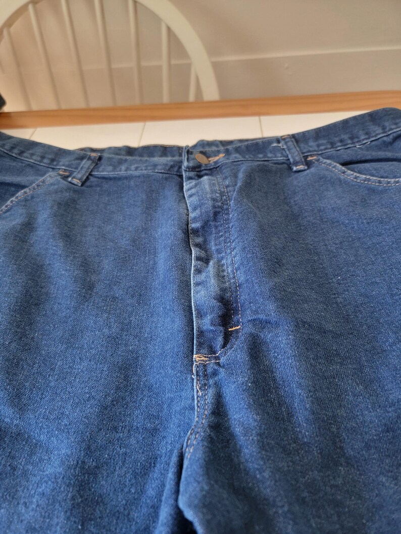 May include: Close-up of blue denim jeans. The jeans have a button closure and pockets. The stitching is visible in a contrasting color. The fabric appears to be a medium weight denim.