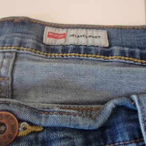 K&ouml;nnte beinhalten: Nahaufnahme einer blauen Jeans mit einem Knopf und einem Etikett mit der Aufschrift "Wrangler RELAXED BOOT". Die Jeans hat gelbe N&auml;hte und ein verwaschenes Aussehen.