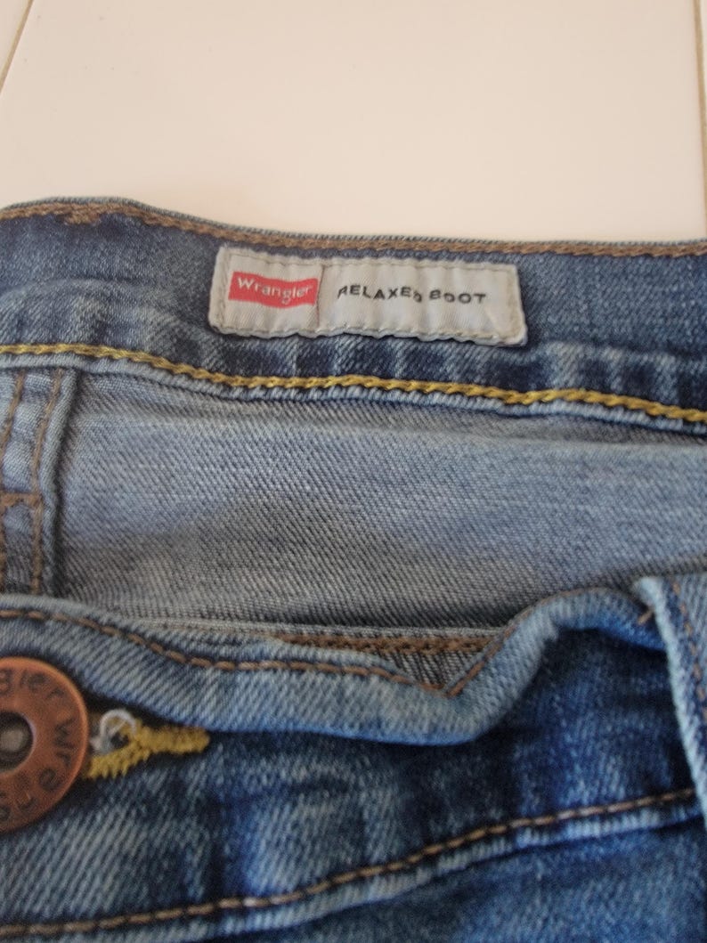 K&ouml;nnte beinhalten: Nahaufnahme einer blauen Jeans mit einem Knopf und einem Etikett mit der Aufschrift "Wrangler RELAXED BOOT". Die Jeans hat gelbe N&auml;hte und ein verwaschenes Aussehen.