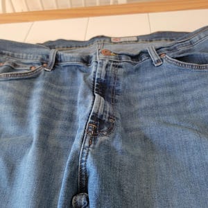 K&ouml;nnte beinhalten: Nahaufnahme einer blauen Jeans. Die Jeans hat einen Knopfverschluss, G&uuml;rtelschlaufen und Taschen. Der Denim hat ein verwaschenes Aussehen mit helleren Bereichen an den Oberschenkeln und Knien. Die Jeans ist f&uuml;r den Alltag geeignet.