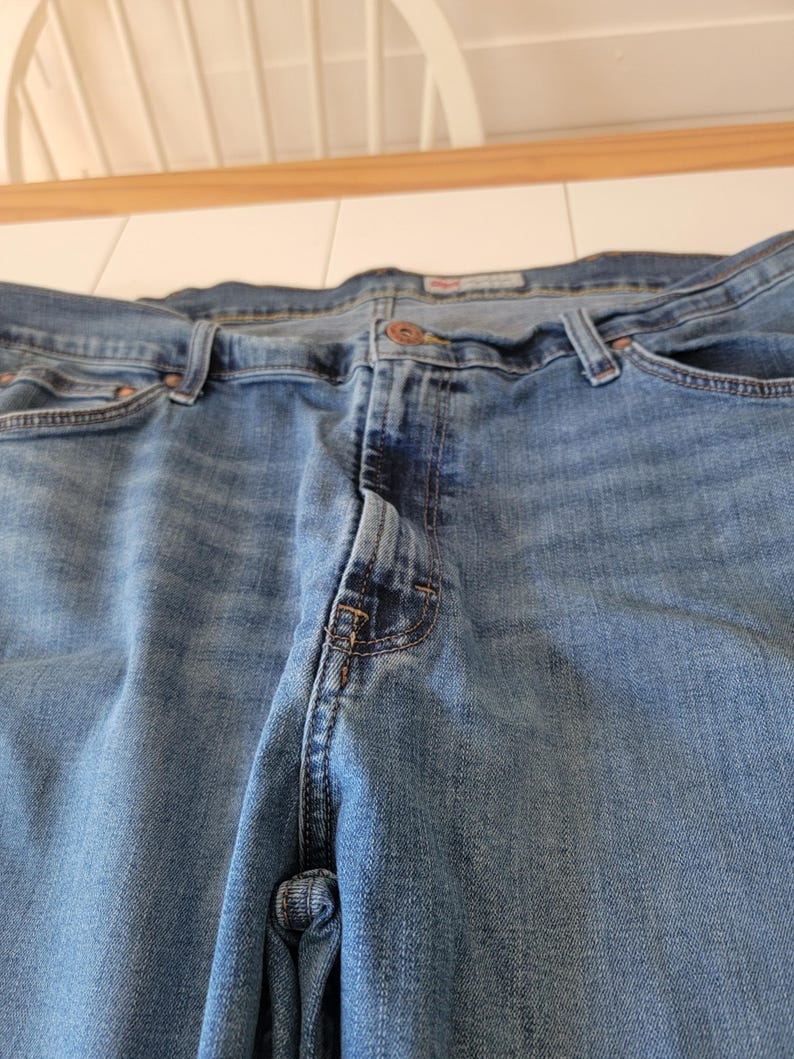 K&ouml;nnte beinhalten: Nahaufnahme einer blauen Jeans. Die Jeans hat einen Knopfverschluss, G&uuml;rtelschlaufen und Taschen. Der Denim hat ein verwaschenes Aussehen mit helleren Bereichen an den Oberschenkeln und Knien. Die Jeans ist f&uuml;r den Alltag geeignet.