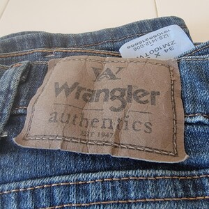 May include: Close-up of blue denim jeans with a brown leather patch featuring the "Wrangler authentics" logo. The jeans have visible stitching and a tag with size and style information.