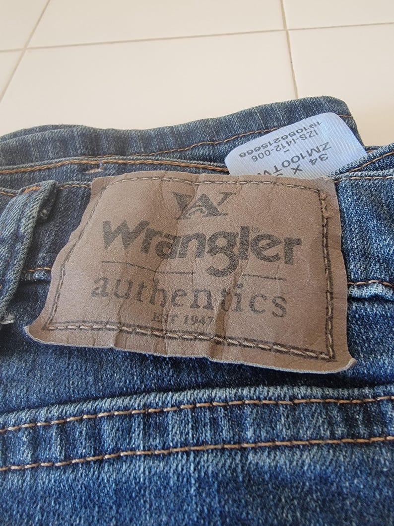 May include: Close-up of blue denim jeans with a brown leather patch featuring the "Wrangler authentics" logo. The jeans have visible stitching and a tag with size and style information.