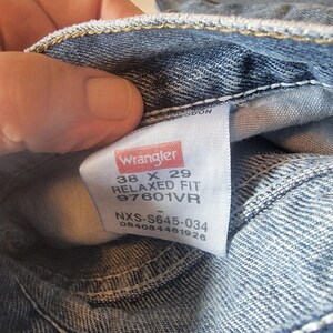 May include: Close-up of blue denim jeans with a white tag. The tag reads "Wrangler" in red and white, with size details: 38 x 29, Relaxed Fit, and style number 97601VR. The jeans are folded.
