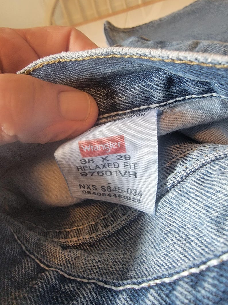 May include: Close-up of blue denim jeans with a white tag. The tag reads "Wrangler" in red and white, with size details: 38 x 29, Relaxed Fit, and style number 97601VR. The jeans are folded.