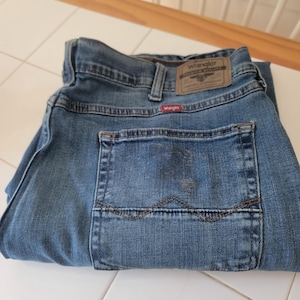 K&ouml;nnte beinhalten: Gefaltete blaue Jeans aus Denim mit dem Wrangler-Markenetikett auf der R&uuml;ckseite. Die Jeans haben ein klassisches F&uuml;nf-Taschen-Design und eine mittelblaue Waschung. Die Ges&auml;&szlig;tasche hat dekorative N&auml;hte.