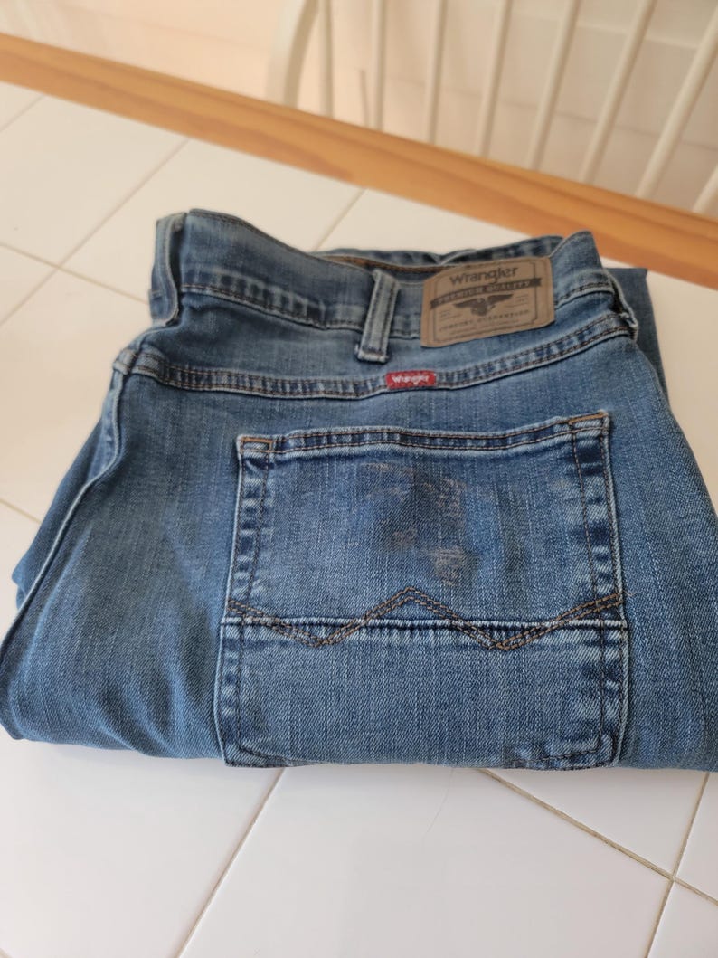 K&ouml;nnte beinhalten: Gefaltete blaue Jeans aus Denim mit dem Wrangler-Markenetikett auf der R&uuml;ckseite. Die Jeans haben ein klassisches F&uuml;nf-Taschen-Design und eine mittelblaue Waschung. Die Ges&auml;&szlig;tasche hat dekorative N&auml;hte.