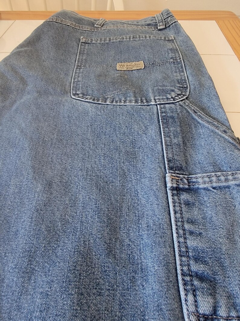 May include: Light blue denim jeans with multiple pockets, including a large carpenter pocket on the side. The back pocket features a small, rectangular "Wrangler" label. The jeans are made of a durable denim fabric.