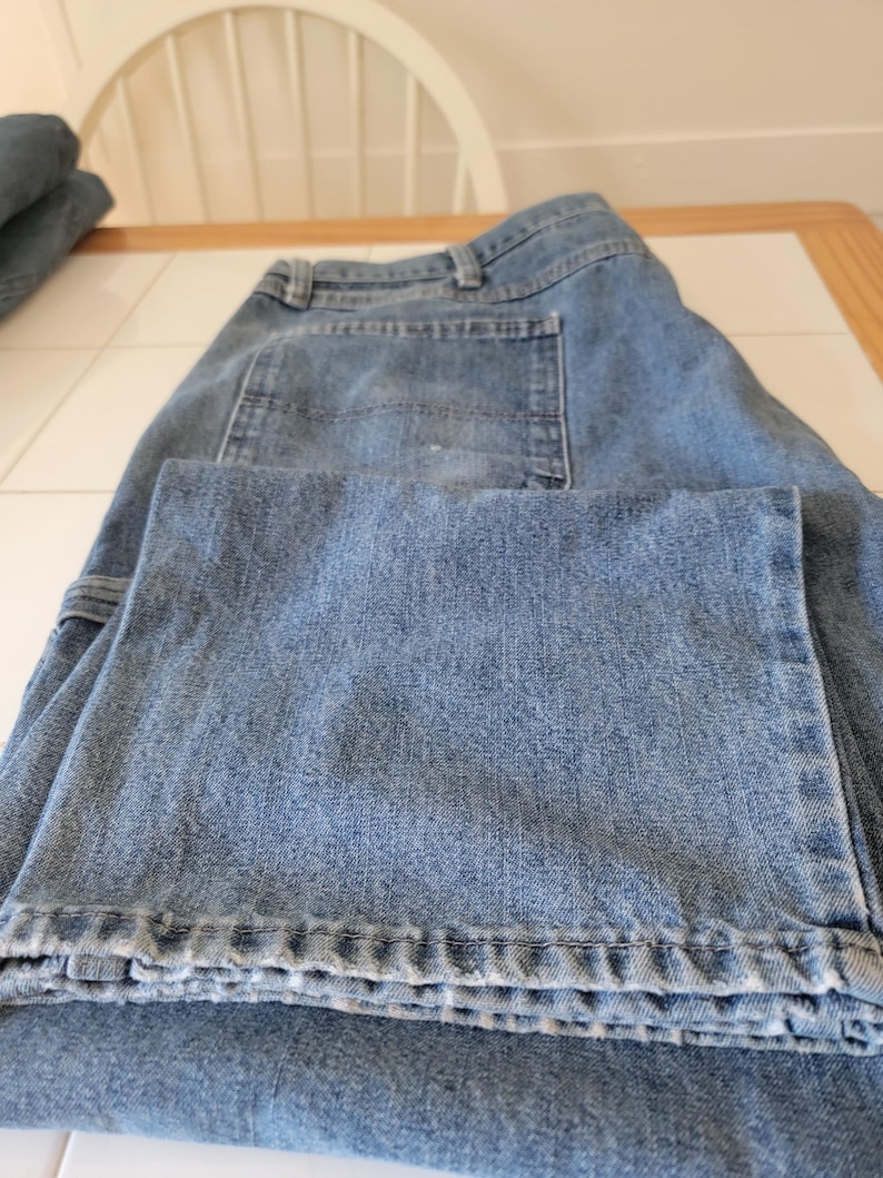 May include: Light blue denim jeans with a classic five-pocket design. The jeans are folded, showcasing the front pockets, belt loops, and the texture of the denim fabric. The jeans appear to be a relaxed fit.