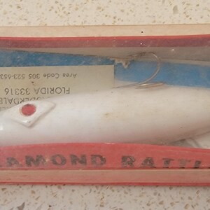May include: A white fishing lure with a red dot on the top, in a clear plastic box with a red and white label that says "Diamond Rattlers". The label also includes the text "Florida 33316,  Overdale,  Area Code 305 553-4331".