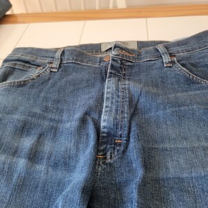 May include: Close-up of blue denim jeans. The jeans have a button and zipper closure, belt loops, and pockets. The denim has a classic, worn look.