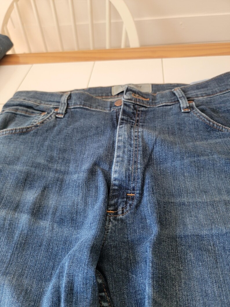 May include: Close-up of blue denim jeans. The jeans have a button and zipper closure, belt loops, and pockets. The denim has a classic, worn look.