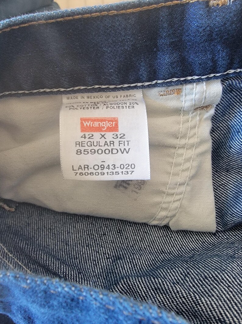 May include: Close-up of a Wrangler jeans label. The label is white with black text, indicating size 42 x 32, regular fit. The jeans are made in Mexico of US fabric, with 80% cotton and 20% polyester.