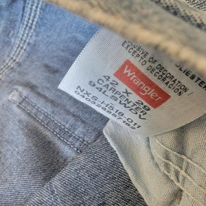 May include: Close-up of a Wrangler jeans tag. The tag is white with red lettering and text that reads "Wrangler 42 X 29 Carpenter 94LSWDV". The jeans are light blue denim.
