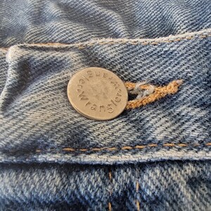 Puede incluir: Primer plano de tela vaquera azul con un bot&oacute;n plateado. El bot&oacute;n est&aacute; grabado con la palabra "Wrangler" en un patr&oacute;n circular. El denim tiene costuras visibles y un aspecto desgastado.