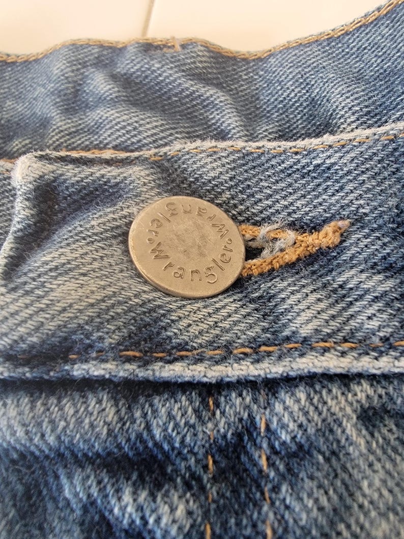 Puede incluir: Primer plano de tela vaquera azul con un bot&oacute;n plateado. El bot&oacute;n est&aacute; grabado con la palabra "Wrangler" en un patr&oacute;n circular. El denim tiene costuras visibles y un aspecto desgastado.