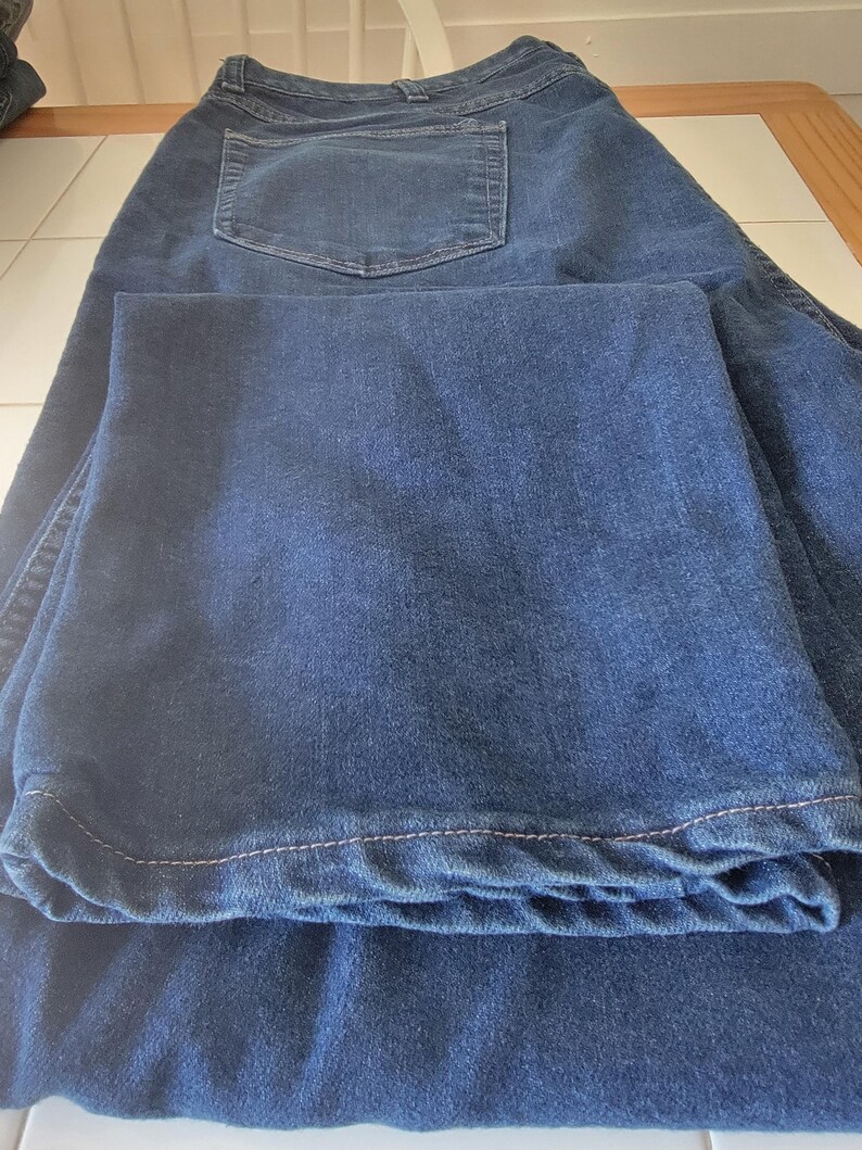 May include: Dark blue denim jeans with a classic five-pocket design. The jeans are folded, showcasing the back pocket and the hem. The fabric appears to be a medium weight, suitable for everyday wear.