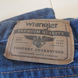 May include: Close-up of a blue denim fabric with a brown leather patch. The patch reads "Wrangler PREMIUM QUALITY" with an eagle emblem and the text "COMFORT. GUARANTEED. GREENSBORO, NORTH CAROLINA."