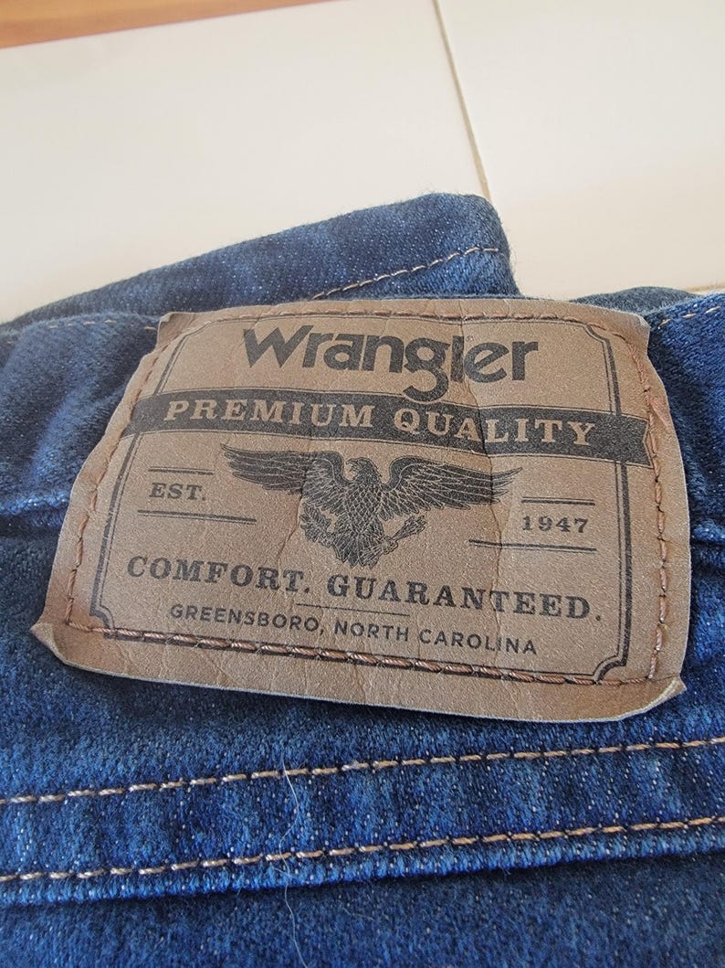 May include: Close-up of a blue denim fabric with a brown leather patch. The patch reads "Wrangler PREMIUM QUALITY" with an eagle emblem and the text "COMFORT. GUARANTEED. GREENSBORO, NORTH CAROLINA."