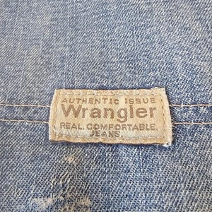 May include: Close-up of blue denim fabric with a rectangular label. The label reads "AUTHENTIC ISSUE Wrangler REAL COMFORTABLE JEANS" in a serif font. The denim has a faded appearance.