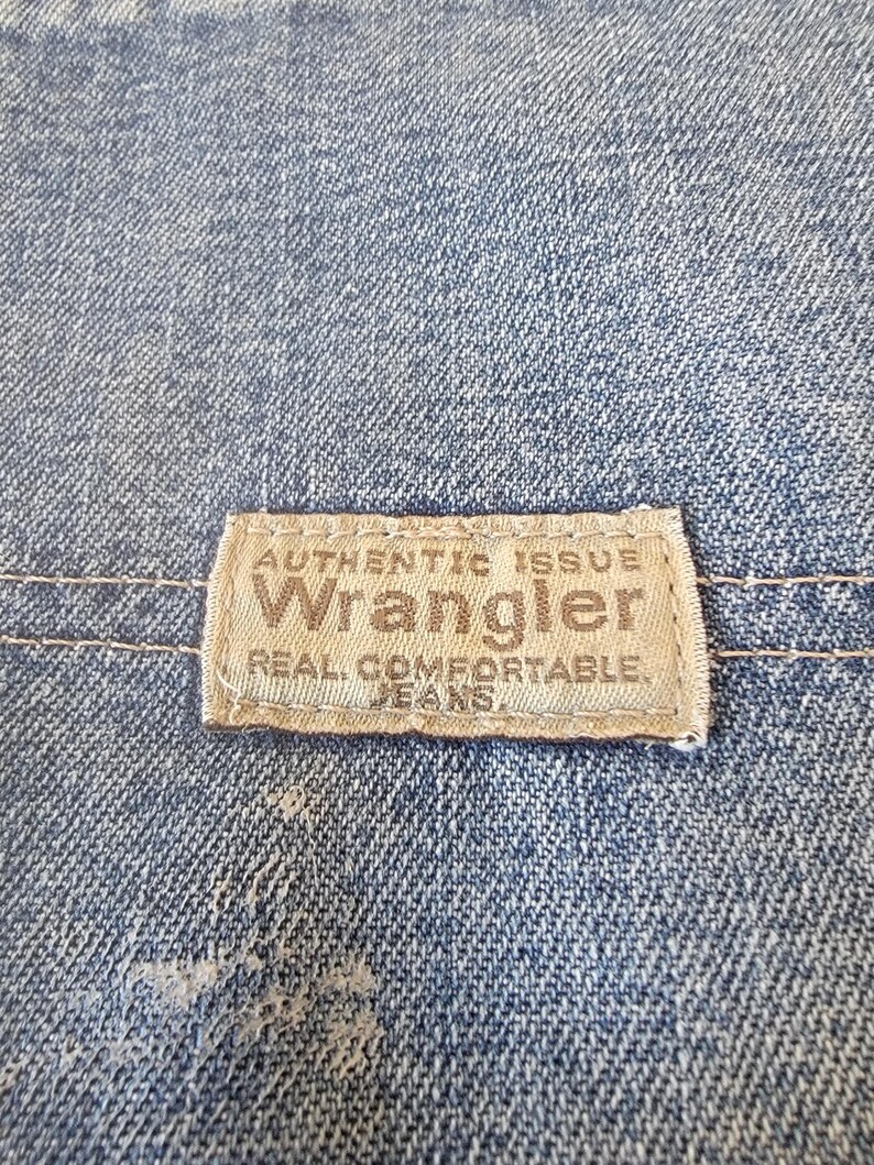 May include: Close-up of blue denim fabric with a rectangular label. The label reads "AUTHENTIC ISSUE Wrangler REAL COMFORTABLE JEANS" in a serif font. The denim has a faded appearance.