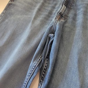 K&ouml;nnte beinhalten: Nahaufnahme einer hellblauen Jeans. Die Jeans hat ein klassisches F&uuml;nf-Taschen-Design, einen Knopf- und Rei&szlig;verschluss und eine verwaschene Optik. Die N&auml;hte sind entlang der N&auml;hte und Taschen sichtbar.
