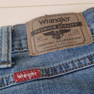 K&ouml;nnte beinhalten: Nahaufnahme einer blauen Jeans mit einem braunen Lederaufn&auml;her. Der Aufn&auml;her tr&auml;gt die Aufschrift "Wrangler PREMIUM QUALITY" mit einem Adleremblem und "COMFORT. GUARANTEED." Das rote Wrangler-Etikett ist sichtbar.