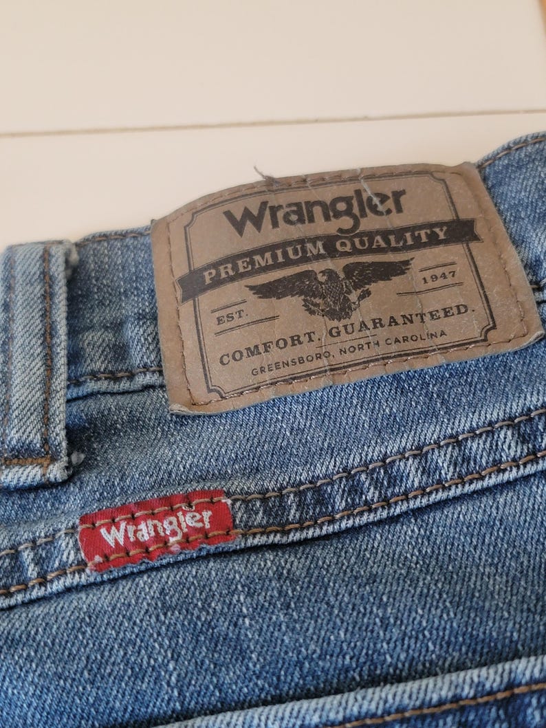K&ouml;nnte beinhalten: Nahaufnahme einer blauen Jeans mit einem braunen Lederaufn&auml;her. Der Aufn&auml;her tr&auml;gt die Aufschrift "Wrangler PREMIUM QUALITY" mit einem Adleremblem und "COMFORT. GUARANTEED." Das rote Wrangler-Etikett ist sichtbar.