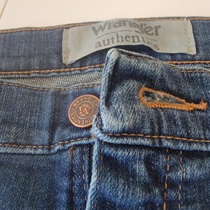 May include: Close-up of blue denim jeans. The image shows the button and zipper area, with the brand name "Wrangler authentics" visible on a label. The jeans have copper-colored stitching and a copper button.