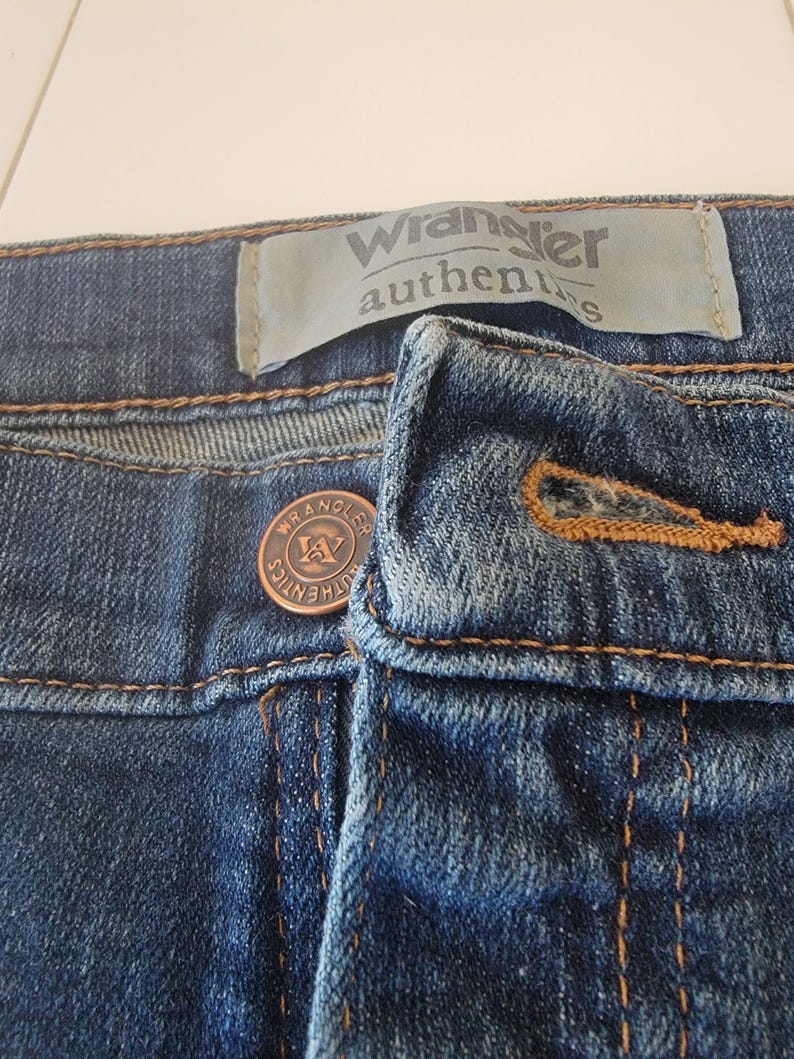 May include: Close-up of blue denim jeans. The image shows the button and zipper area, with the brand name "Wrangler authentics" visible on a label. The jeans have copper-colored stitching and a copper button.