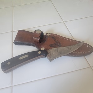 May include: A fixed-blade knife with a dark handle and a metallic blade featuring the text SCHRADE USA 152. The knife rests on a brown leather sheath with a snap closure and an embossed eagle design, featuring visible stitching along the edges.