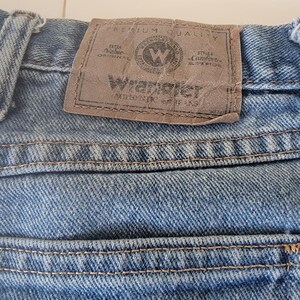 Puede incluir: Primer plano de unos vaqueros de mezclilla azules con un parche de cuero marr&oacute;n. El parche presenta el logotipo de Wrangler y el texto "Premium Quality" y "Extra Value Original".