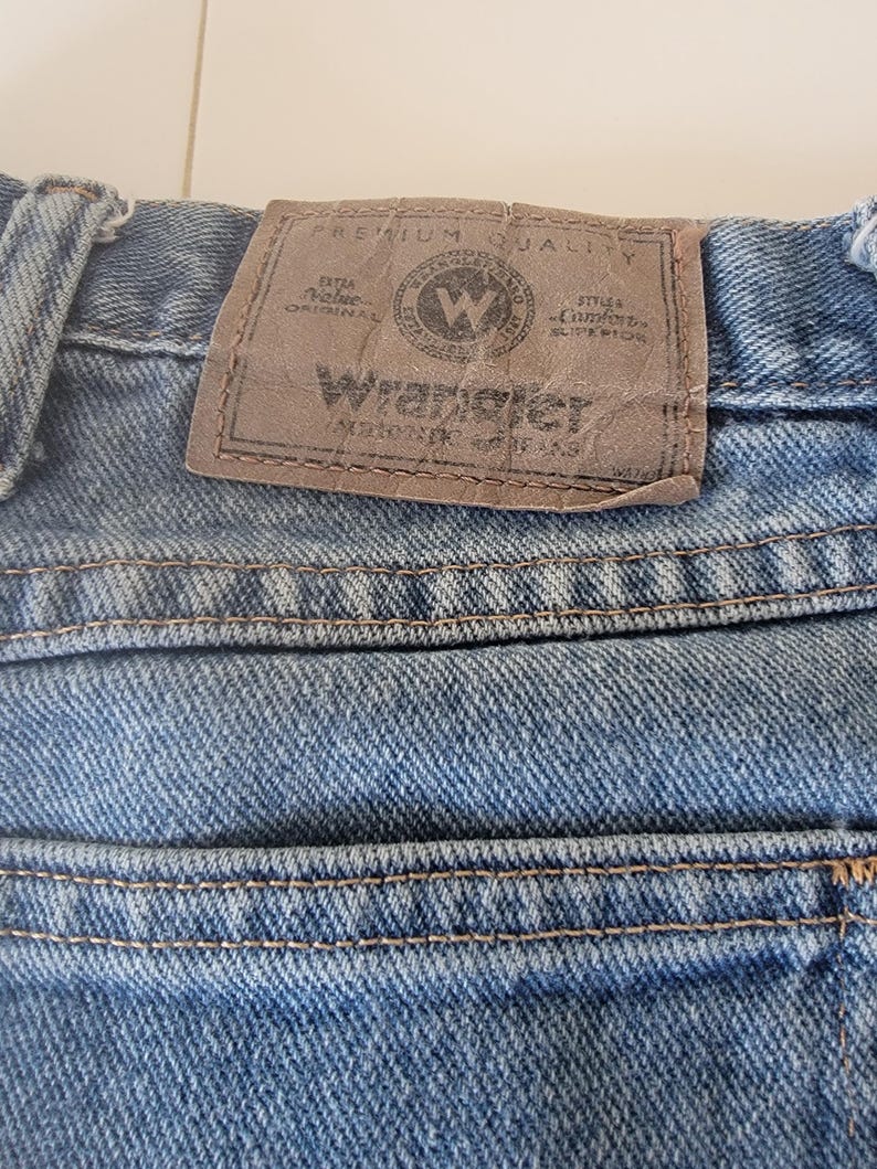 Puede incluir: Primer plano de unos vaqueros de mezclilla azules con un parche de cuero marr&oacute;n. El parche presenta el logotipo de Wrangler y el texto "Premium Quality" y "Extra Value Original".