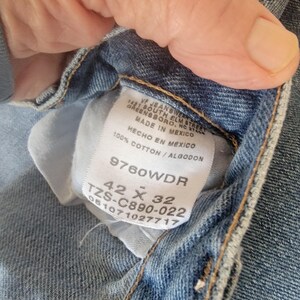 Puede incluir: Primer plano de una etiqueta de vaqueros de mezclilla azul claro. La etiqueta dice "VF Jeanswear, Greensboro, NC, Made in Mexico, 100% Cotton". Los vaqueros son de talla 42 x 32.