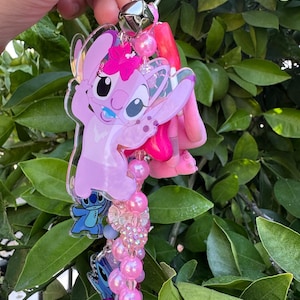 May include: A keychain featuring a large pink acrylic character charm with a flower, smaller blue character charms, a silver metal bell, and a strand of pink, purple, and clear beads in various textures including smooth, faceted, and glittery finishes.