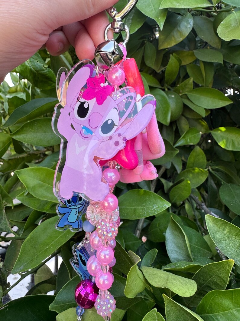 May include: A keychain featuring a large pink acrylic character charm with a flower, smaller blue character charms, a silver metal bell, and a strand of pink, purple, and clear beads in various textures including smooth, faceted, and glittery finishes.