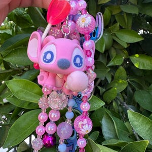 May include: A decorative keychain featuring a pink character figure with large blue eyes, accented by a bright pink lollipop charm and a variety of pink, purple, and light blue beads. The design includes floral-patterned beads and clear acrylic charms.
