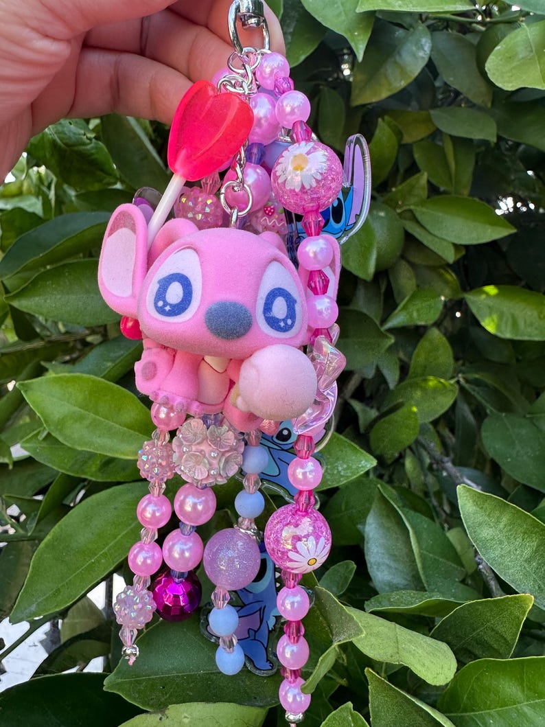 May include: A decorative keychain featuring a pink character figure with large blue eyes, accented by a bright pink lollipop charm and a variety of pink, purple, and light blue beads. The design includes floral-patterned beads and clear acrylic charms.