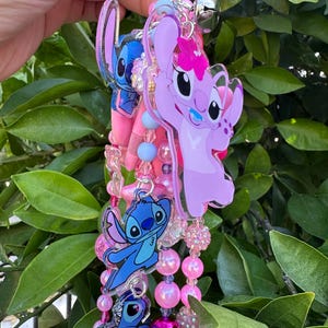 May include: A decorative keychain featuring multiple acrylic charms of blue and pink cartoon characters, including a pink character with a flower, interspersed with various pink, blue, and clear beads, a silver metal clasp, and a small silver bell.