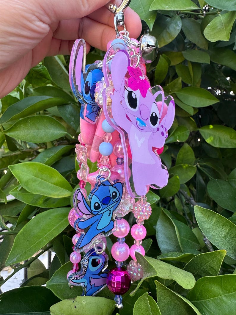 May include: A decorative keychain featuring multiple acrylic charms of blue and pink cartoon characters, including a pink character with a flower, interspersed with various pink, blue, and clear beads, a silver metal clasp, and a small silver bell.