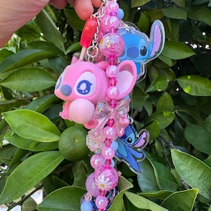 May include: A keychain featuring a pink character figure, two blue character charms, and a strand of pink and blue beads. The beads include clear, glittery, and daisy-patterned spheres, along with a translucent bow charm and a small red popsicle charm.