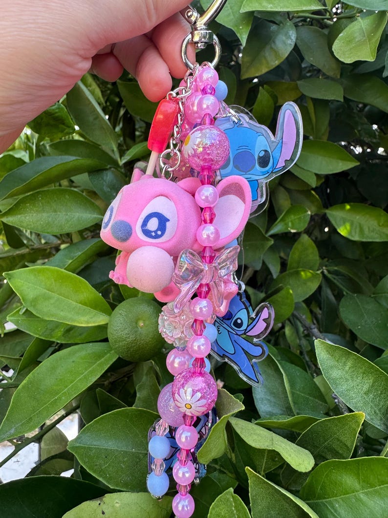 May include: A keychain featuring a pink character figure, two blue character charms, and a strand of pink and blue beads. The beads include clear, glittery, and daisy-patterned spheres, along with a translucent bow charm and a small red popsicle charm.